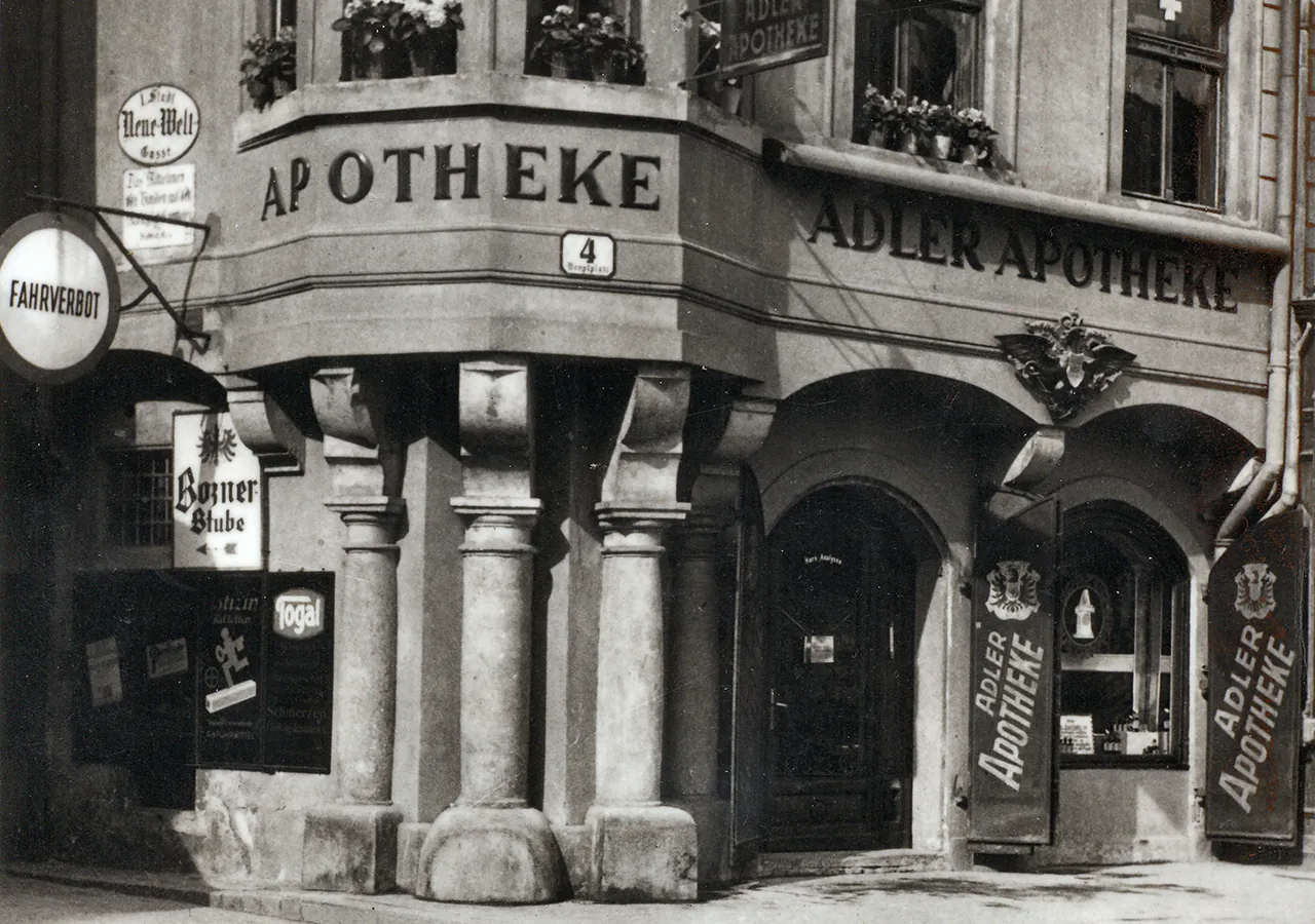 Historisches Schwarz-Weiß-Foto der Fassade der Adler Apotheke aus den 1930er-Jahren mit Säulen und Werbeschildern