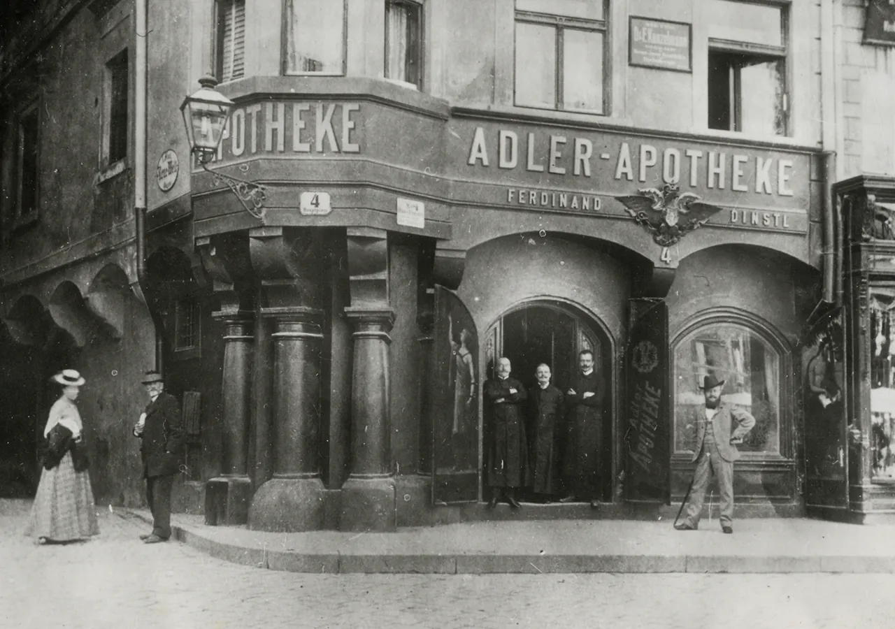 Historisches Schwarz-Weiß-Foto der Adler Apotheke um 1900 mit dem Schild „ADLER-APOTHEKE“ und Passanten vor dem Eingang