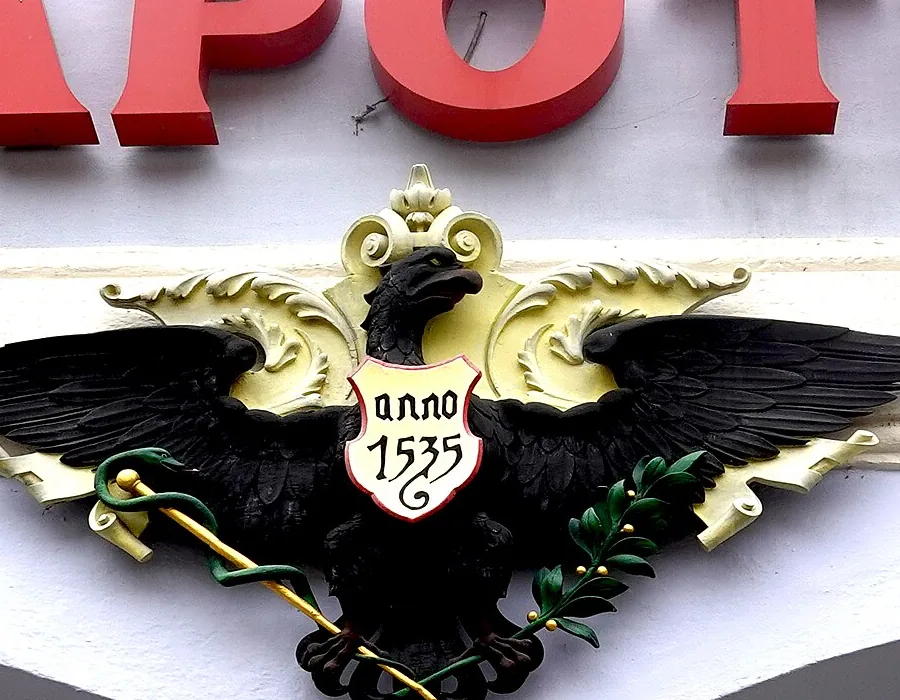 Schwarzer Adler mit goldener Krone und weißem Schild mit der Aufschrift „anno 1535“ an der Fassade der Adler Apotheke