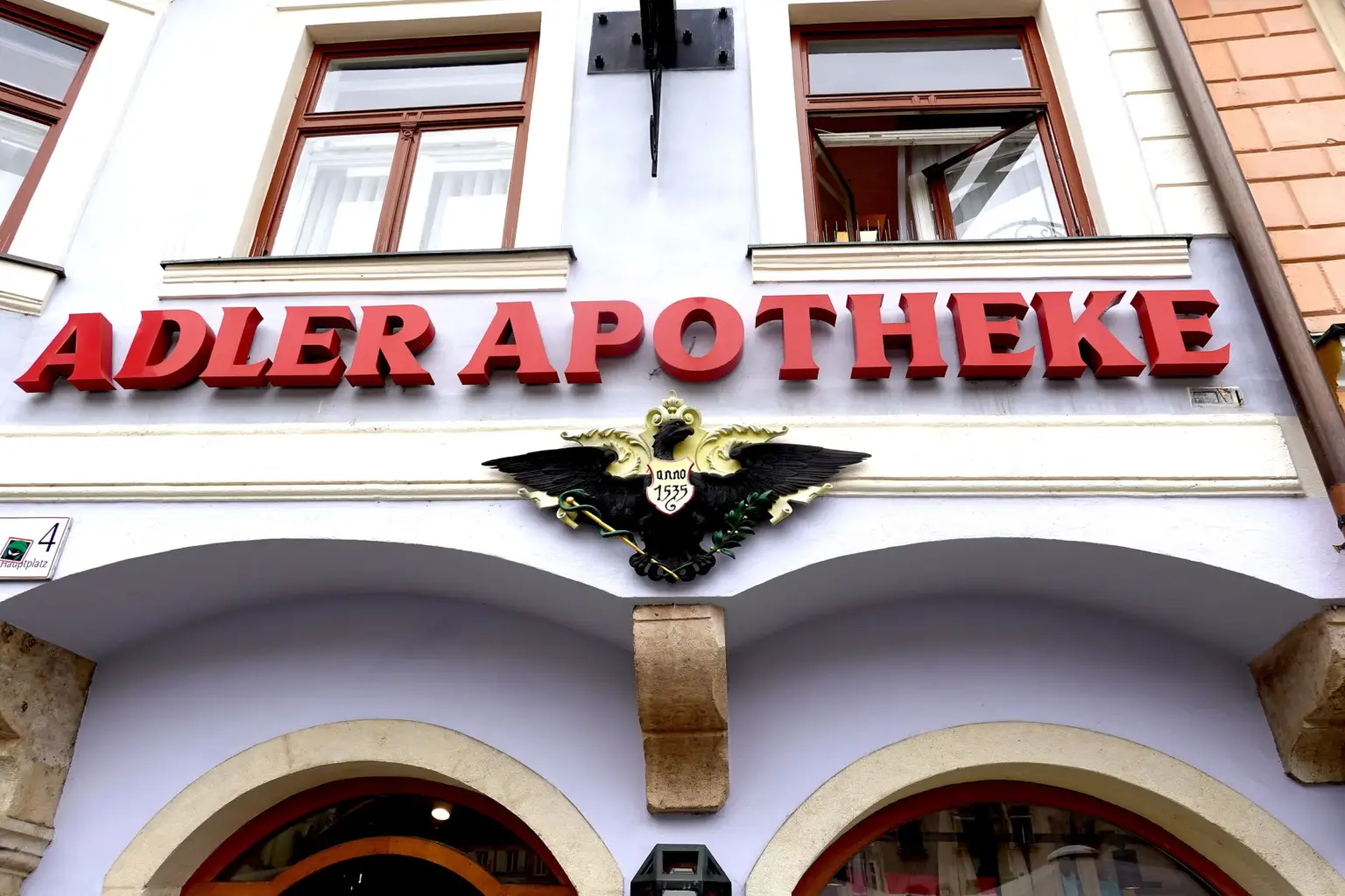Fassade der Adler Apotheke am Grazer Hauptplatz mit rotem Schriftzug „ADLER APOTHEKE“ und dem schwarz-goldenen Adler-Emblem „anno 1535“ über den Arkaden